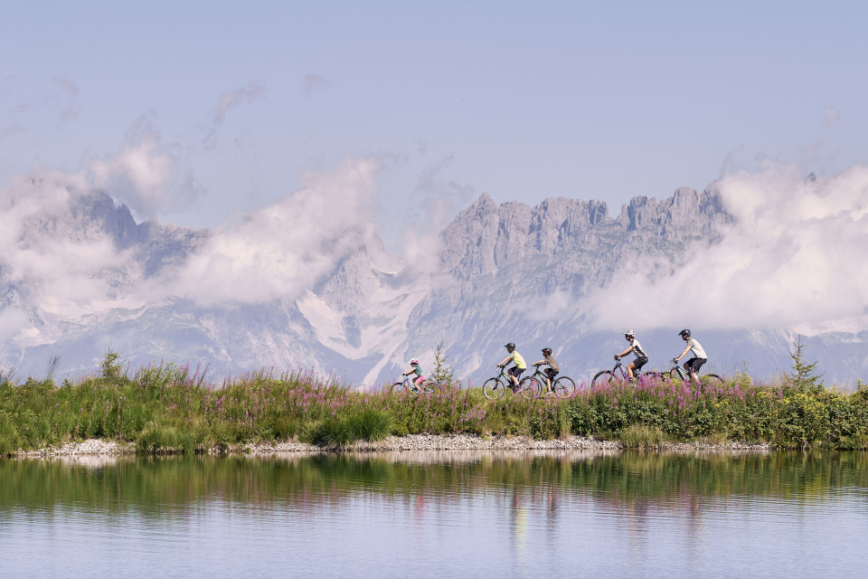Drei Radfahrer fahren auf einem kurvenreichen Weg durch eine üppig grüne Berglandschaft bergauf und vermitteln unter einem teilweise bewölkten Himmel ein Gefühl von Abenteuer und Entschlossenheit.