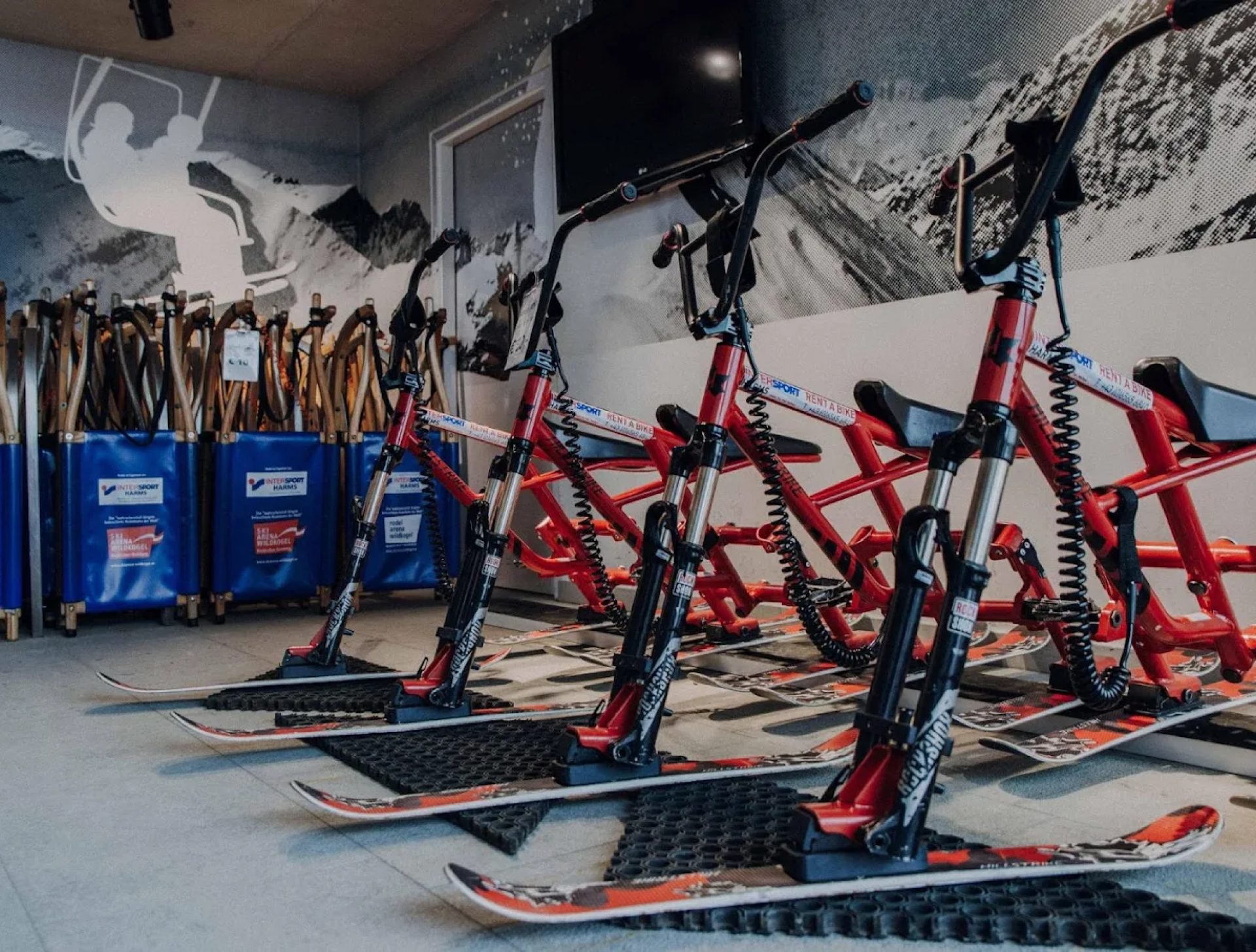Rote Schneemobile mit Monoski-Untergestellen stehen drinnen vor einer Wandmalerei mit schneebedeckten Bergen aufgereiht und schaffen eine abenteuerliche und dynamische Atmosphäre.