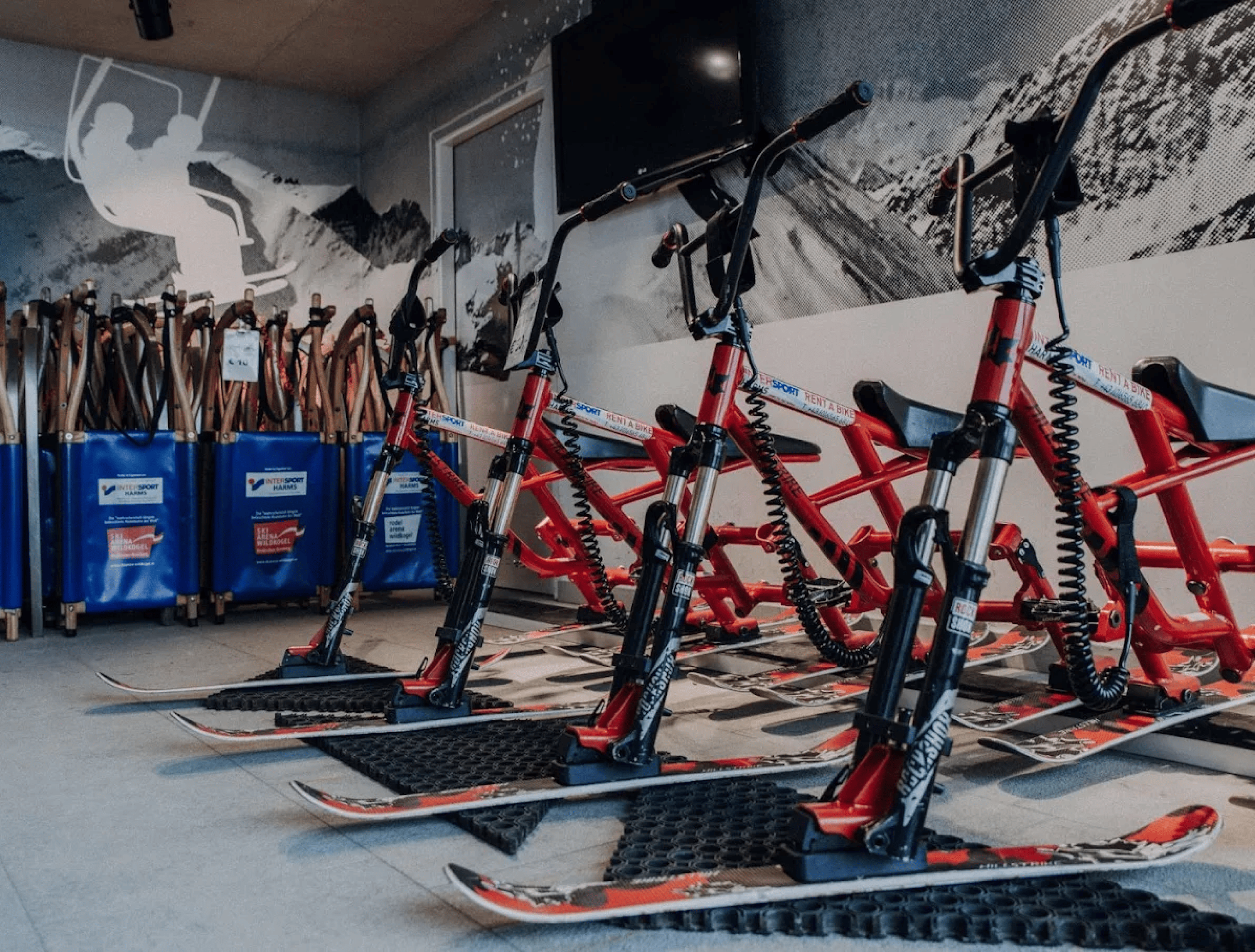 Red snowmobiles with monoski undercarriages are lined up indoors in front of a mural depicting snow-covered mountains, creating an adventurous and dynamic atmosphere.