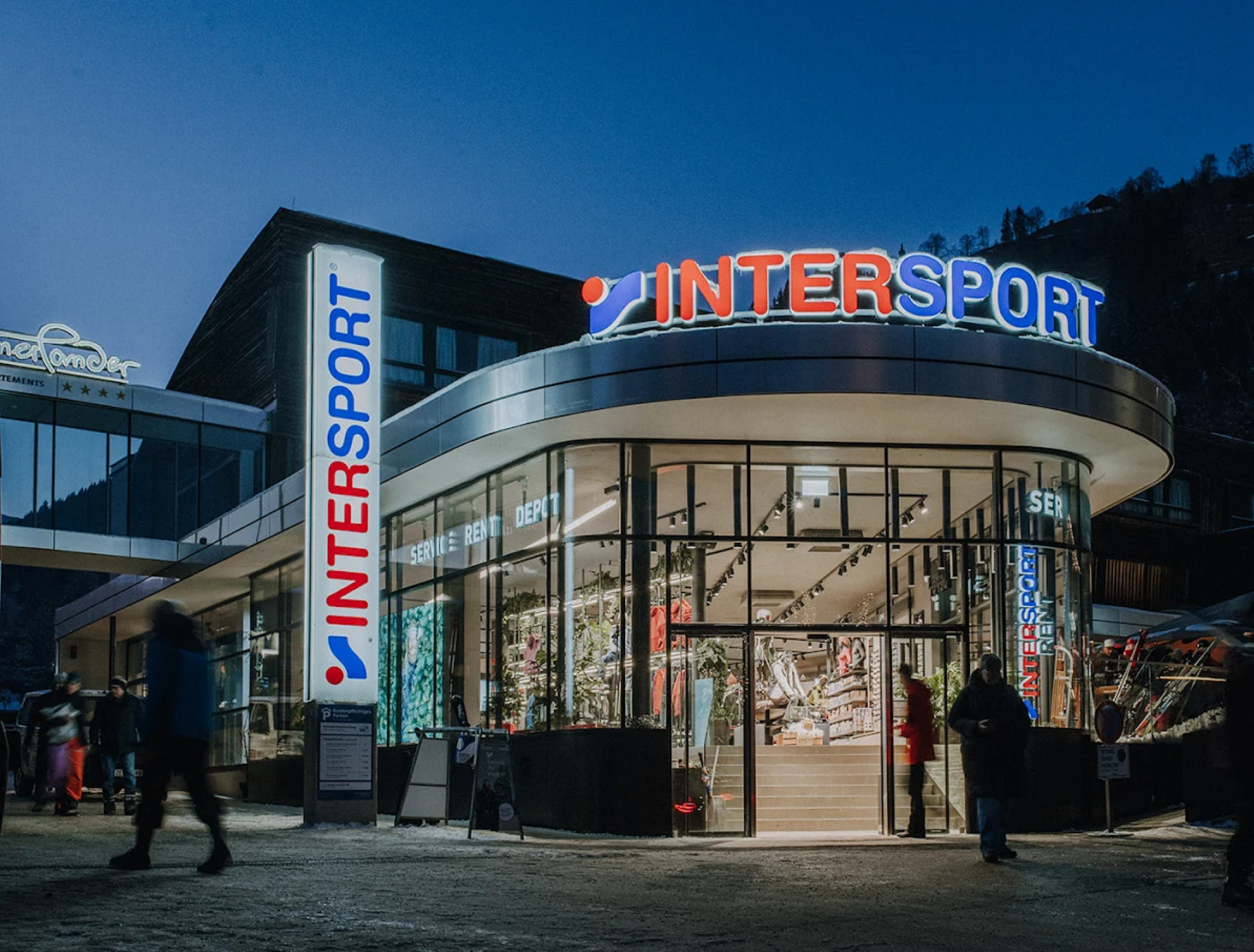 Außenansicht eines Intersport-Geschäfts mit heller Beschilderung in der Abenddämmerung. Menschen gehen vorbei, die geschwungene Glasfassade gibt den Blick auf Sportartikel im Inneren frei. Die Szene ist lebhaft.