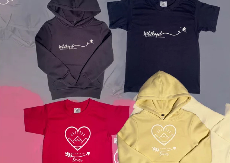 Es werden vier Shirts gezeigt: ein grauer und ein schwarzer Hoodie sowie ein rotes und ein gelbes T-Shirt. Jedes Shirt ist mit einem Herz- oder Pfeilmotiv verziert, das Wärme vermittelt.