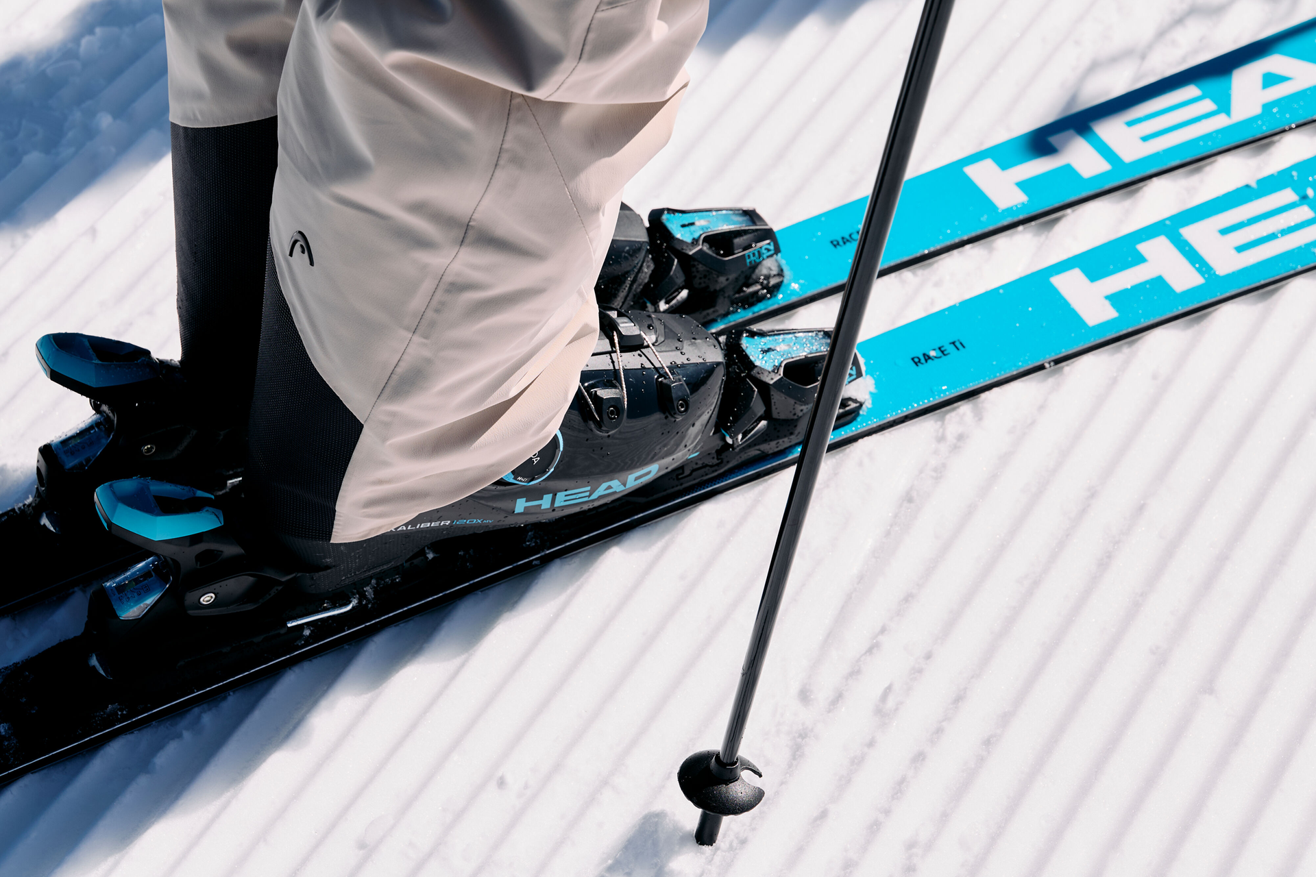 Close-up of a skier's feet on blue HEAD skis and snow, with grooved tracks on a sunny day. Ski poles and beige pants are visible, conveying readiness and focus.