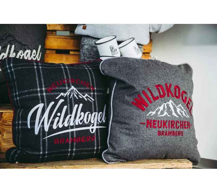 Gemütliche Kissen auf einem Holzregal, eines mit schwarzem Karomuster, das andere grau. Beide sind mit „Wildkogel” und Bergmotiven bedruckt und vermitteln ein rustikales Flair.