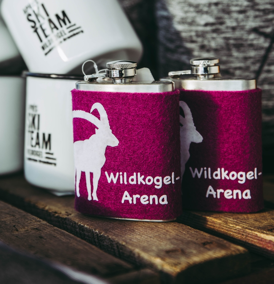 Zwei Edelstahlflaschen, umhüllt von kastanienbraunem Stoff mit einer weißen Ziegensilhouette und dem Schriftzug „Wildkogel-Arena“, auf einer Holzunterlage platziert.