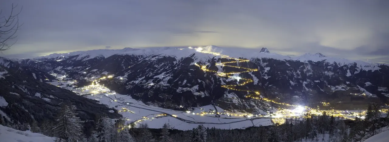 A panoramic night view of a snowy mountain ski resort. Illuminated ski trails snake down dark mountains under a cloudy sky, creating a serene, glowing scene.