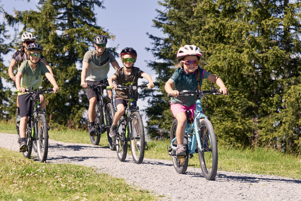Eine Gruppe von drei Kindern und zwei Erwachsenen fährt mit dem Fahrrad auf einem Schotterweg durch üppig grüne Bäume. Alle tragen Helme und Sonnenbrillen und genießen den sonnigen Tag.