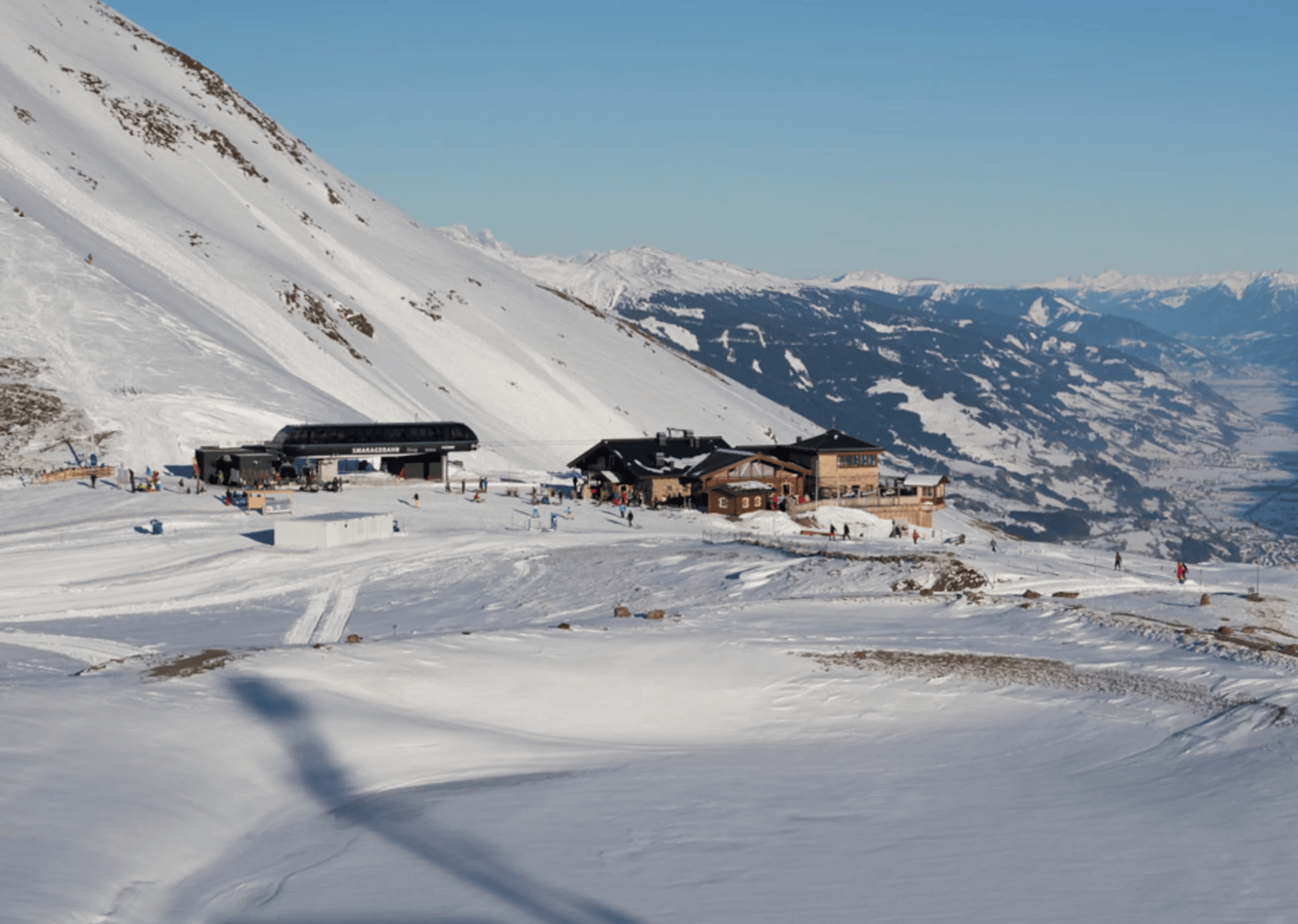 Snowy mountain scene with a ski resort nestled on the slopes, featuring several wooden buildings. Skiers traverse the bright, sunlit snow with mountains in the background.