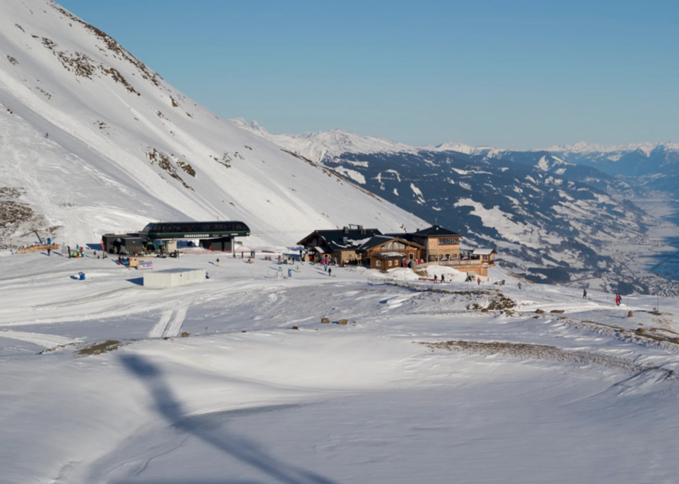 Verschneite Berglandschaft mit einem Skigebiet, das sich an die Hänge schmiegt, mit mehreren Holzgebäuden. Skifahrer gleiten über den hellen, sonnenbeschienenen Schnee, im Hintergrund sind Berge zu sehen.