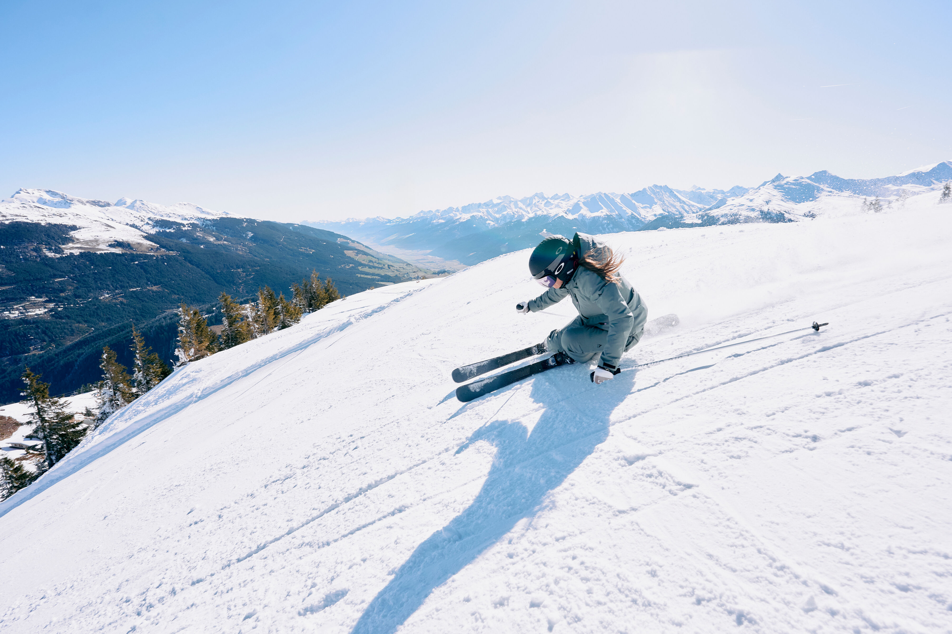 A skier in green gear swiftly descends a snowy slope under a clear blue sky, with distant snow-capped mountains and a valley in the background.