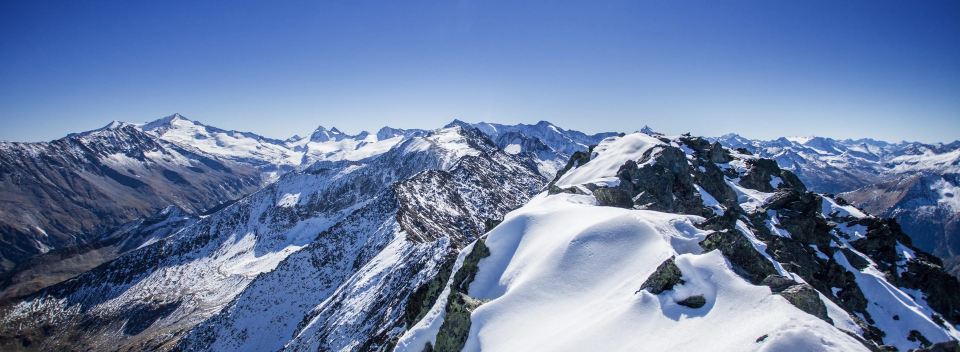 Schneebedeckte Berge unter einem klaren blauen Himmel, die eine ruhige und majestätische Atmosphäre vermitteln. Zackige Gipfel erstrecken sich bis zum Horizont.