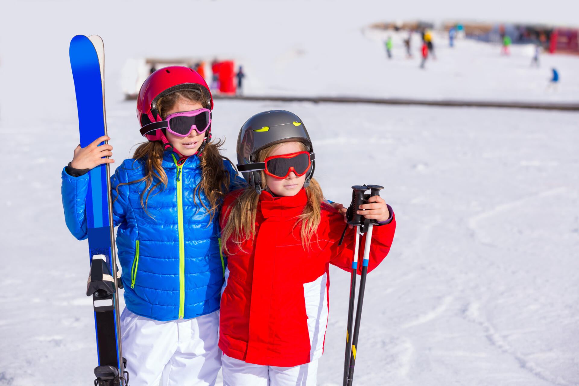 Two children in ski gear stand on a snowy slope. One holds skis and wears a blue jacket; the other holds poles, in a red jacket. They appear cheerful.