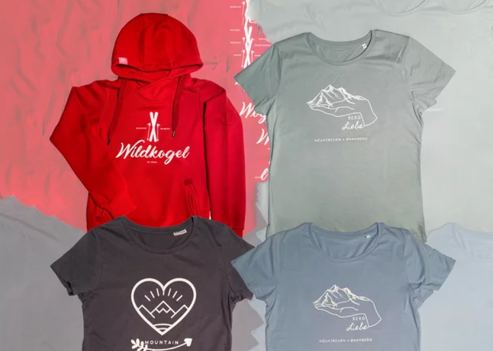 A red hoodie with "Wildkogel" text and mountain logo, surrounded by four gray T-shirts featuring nature-themed designs, arranged on a textured background.