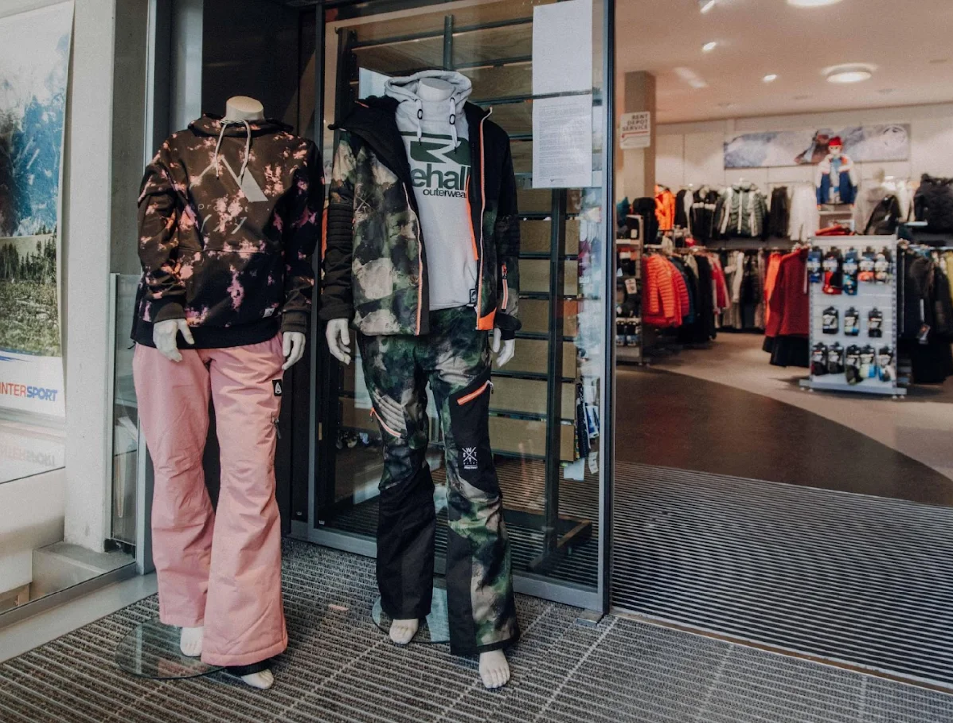 Zwei Schaufensterpuppen in Wintersportbekleidung stehen am Eingang eines Geschäfts. Die eine trägt eine geblümte Jacke und eine rosa Hose, die andere eine Jacke und Hose in Camouflage-Optik. Im Inneren sind weitere Kleidungsstücke ausgestellt.