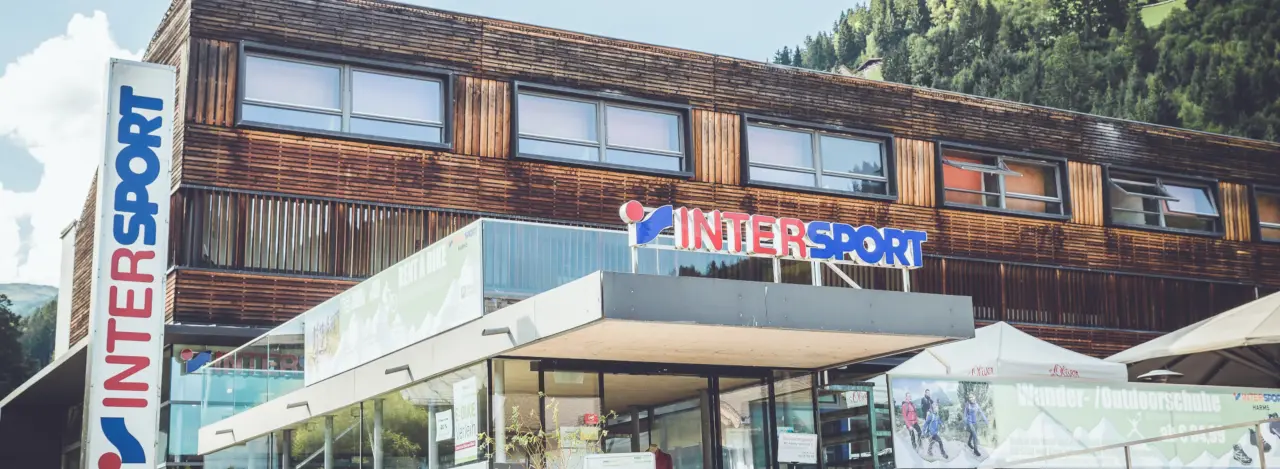 Ein modernes Sportgeschäft mit einer Holzfassade und großen Fenstern. Das „INTERSPORT“-Logo ist gut sichtbar angebracht. Die Szene ist hell und sonnig.