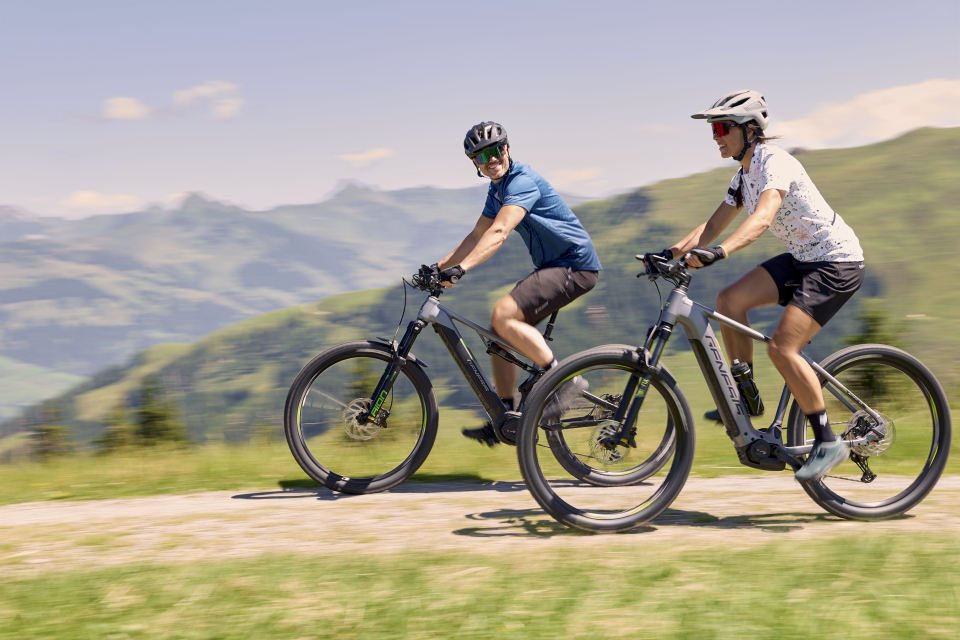 Zwei Radfahrer fahren an einem sonnigen Tag mit Mountainbikes, tragen Helme und Sportbekleidung. Sie befinden sich auf einem Feldweg mit grünen Hügeln und Bergen im Hintergrund.