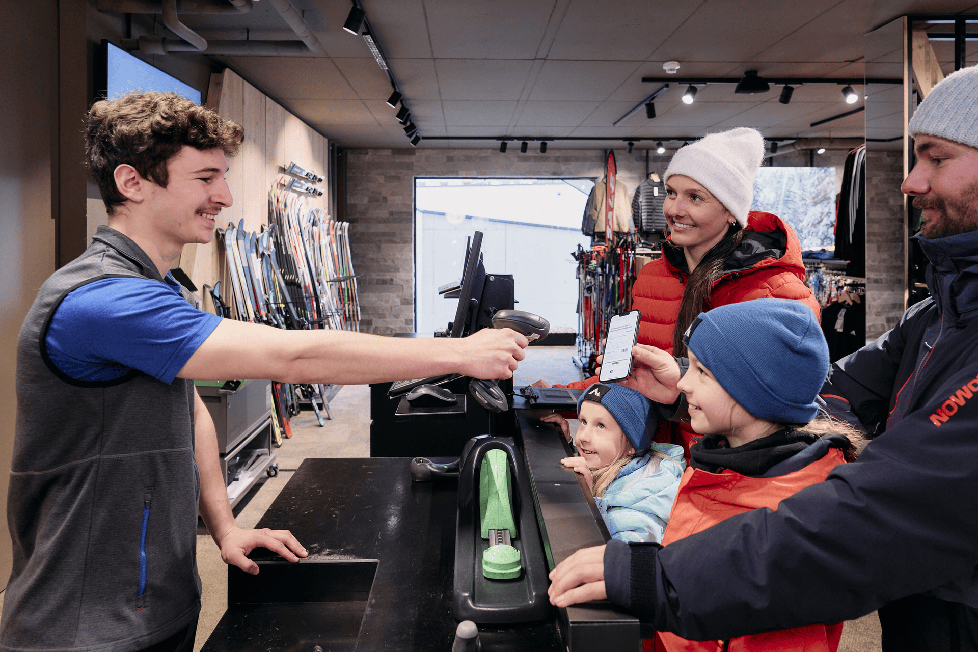 Ein lächelnder Verkäufer scannt das Smartphone einer Familie an der Kasse eines Skigeschäfts. Die Familie, gekleidet in Winterkleidung, wirkt glücklich und aufgeregt. Im Hintergrund sind Skier ausgestellt.