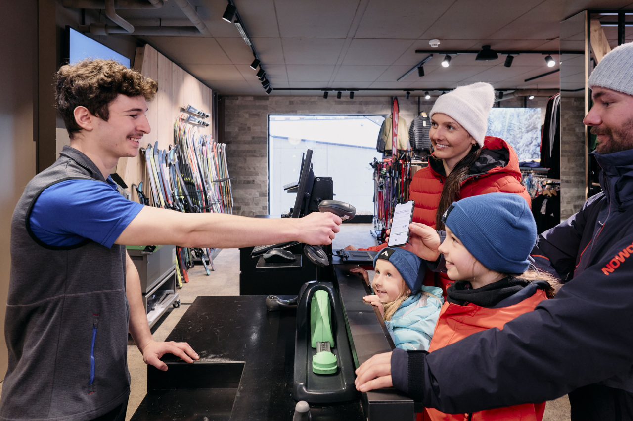 Ein lächelnder Verkäufer scannt das Smartphone einer Familie an der Kasse eines Skigeschäfts. Die Familie, gekleidet in Winterkleidung, wirkt glücklich und aufgeregt. Im Hintergrund sind Skier ausgestellt.
