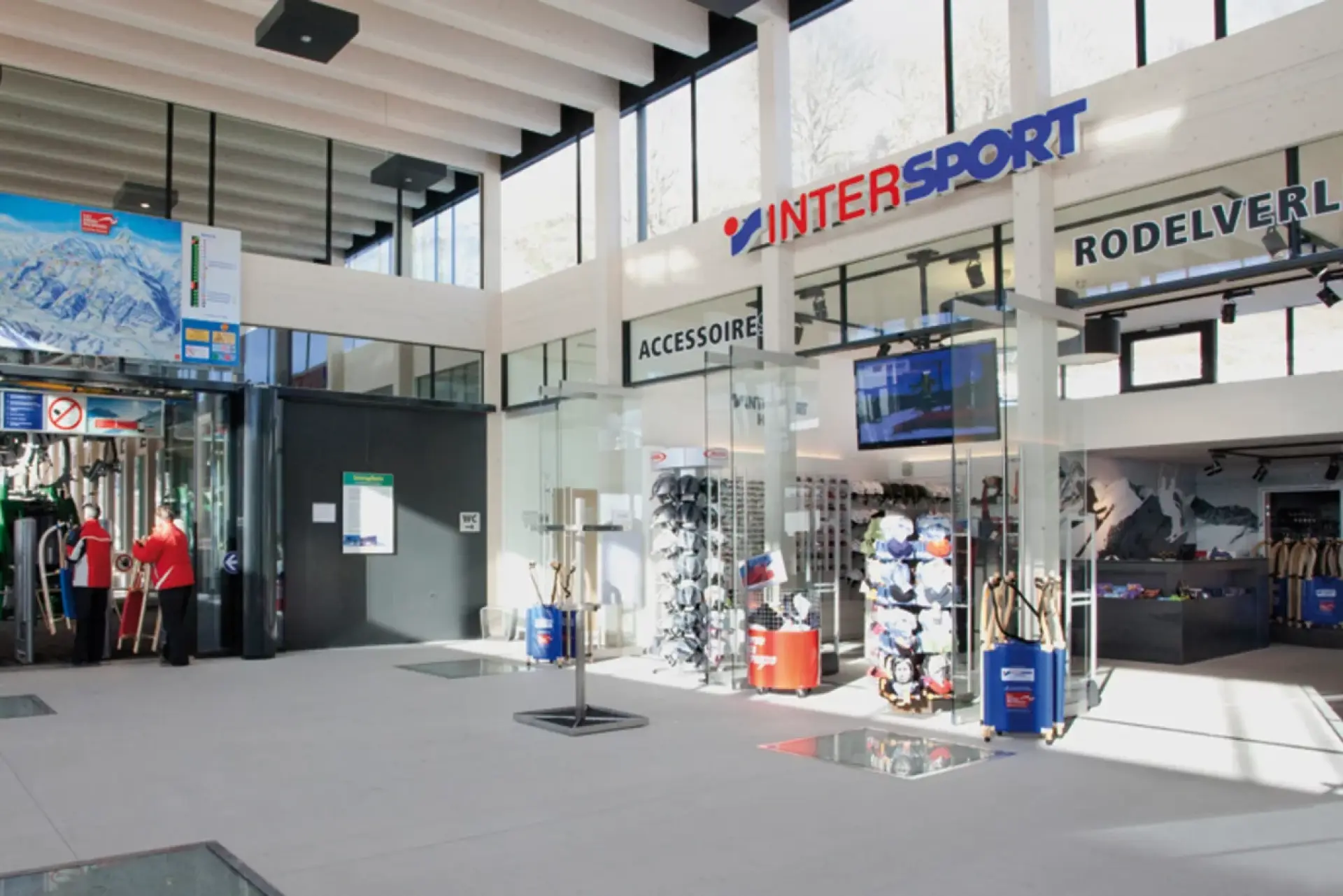 Bright ski rental shop entrance with high windows and modern decor. Two staff members in red jackets provide assistance near skiing gear.