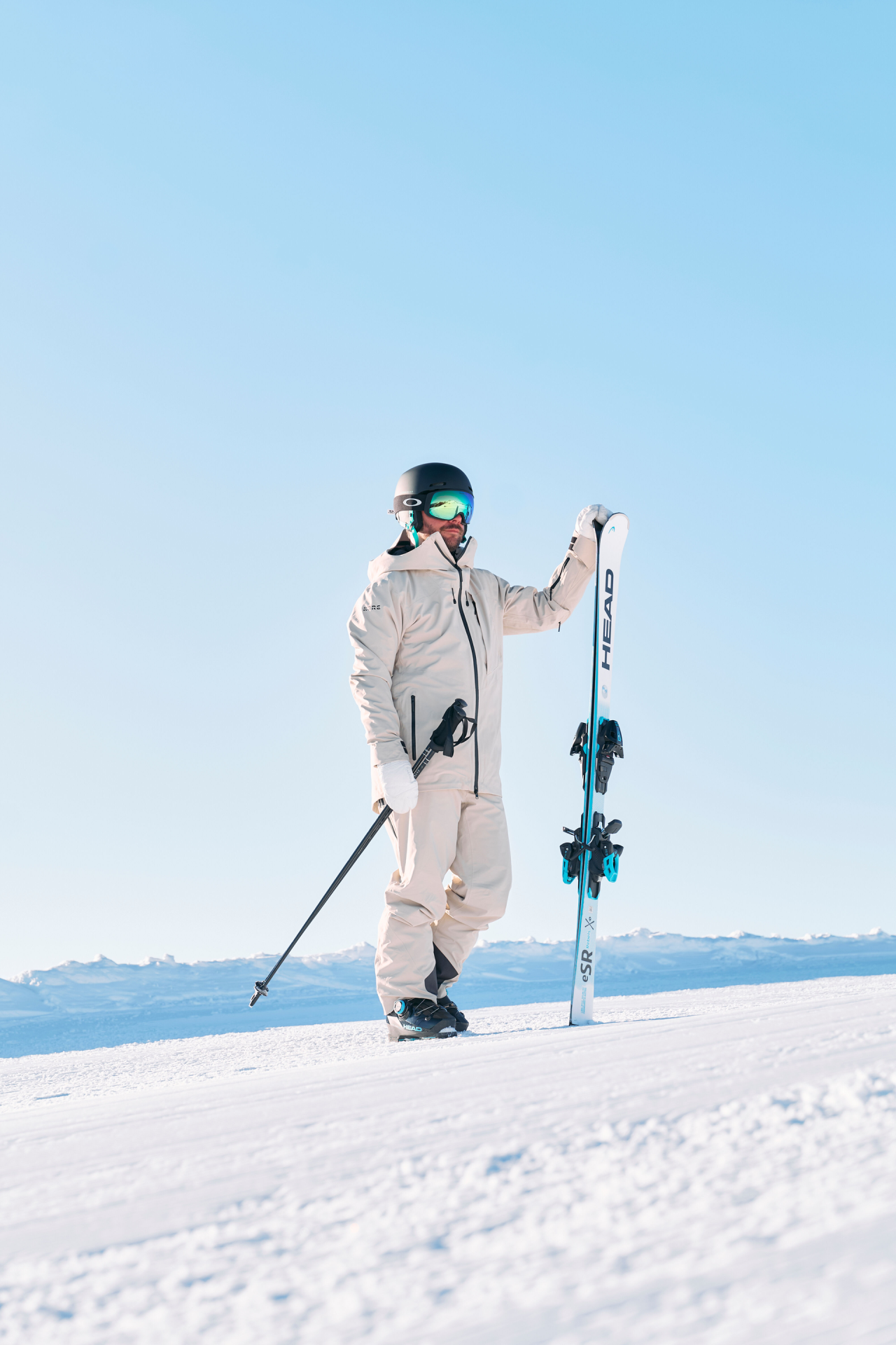 A skier in white gear, helmet, and goggles stands on a snowy slope under a clear blue sky, holding poles and a ski upright, conveying a sense of adventure.