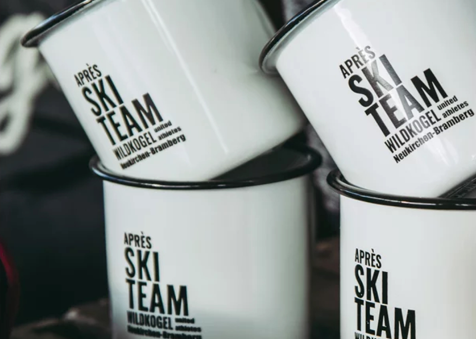 Gestapelte weiße Emaille-Becher mit schwarzem Rand und auffälligem Schriftzug „Après Ski Team Wildkogel“, die eine gemütliche, abenteuerliche Ski-Atmosphäre vermitteln.