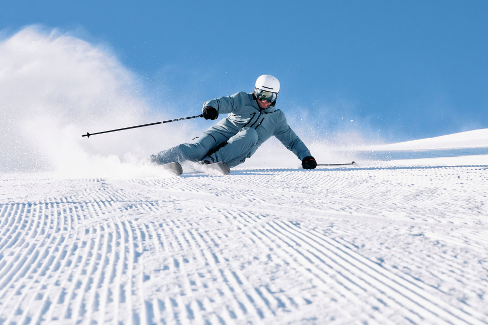 Ein Skifahrer in blauer Kleidung und mit Helm fährt eine sonnige, präparierte Piste hinunter und wirbelt dabei Schnee auf. Die Szene vermittelt Geschwindigkeit und Begeisterung vor einem klaren blauen Himmel.