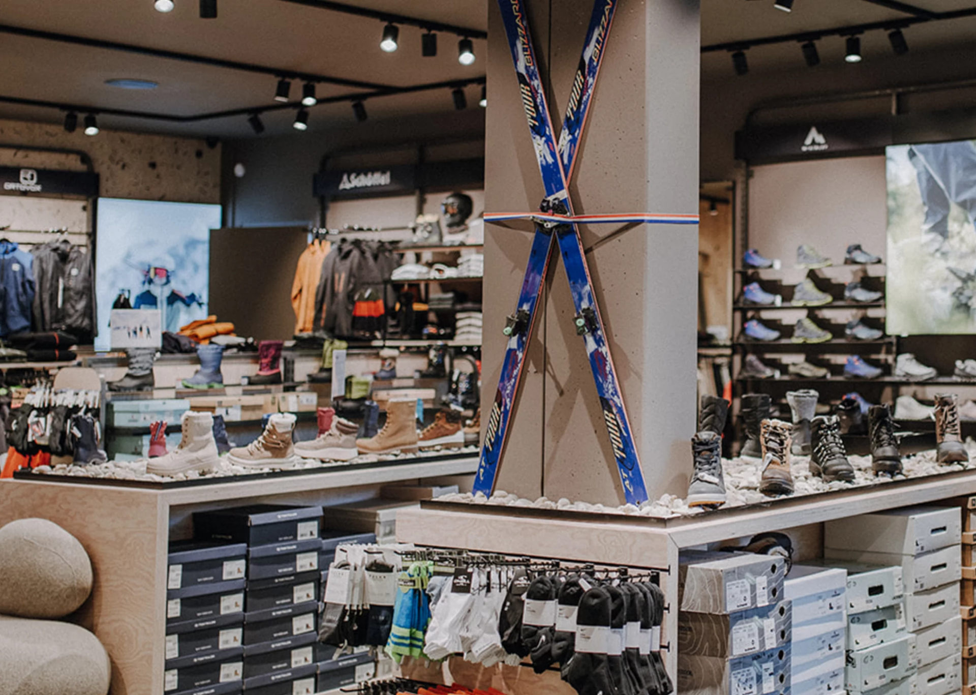 A well-lit outdoor gear store with skis mounted on a pillar, surrounded by hiking boots, boxes, and various outdoor apparel, conveying an adventurous tone.