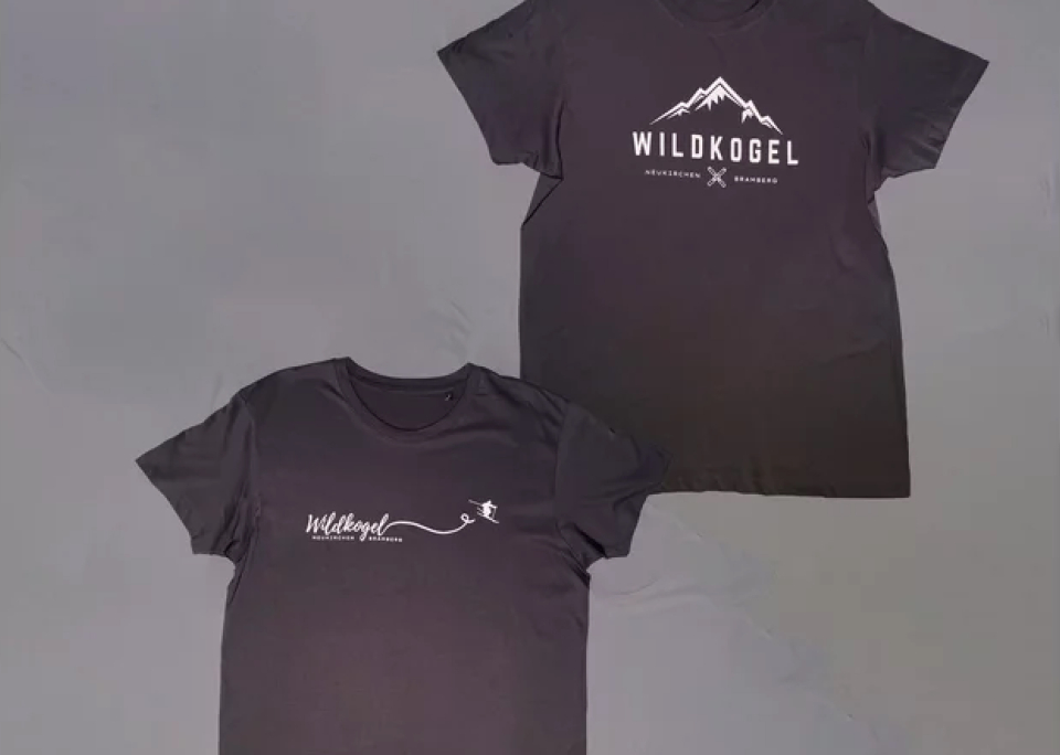 Two black T-shirts feature "Wildkogel" in white. The top shirt has a mountain graphic; the bottom shows a flowing line with a star. Simple and outdoor-inspired.