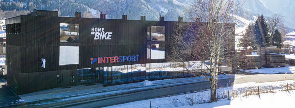 Ein modernes, dunkles Gebäude mit den Aufschriften „Home of Bike“ und „INTERSPORT“ in einer verschneiten Landschaft, mit Bergen im Hintergrund unter einem klaren Himmel.