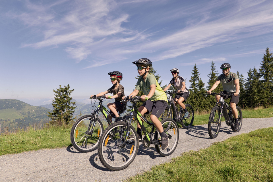 Eine vierköpfige Familie genießt eine Radtour auf einem Schotterweg durch eine malerische Landschaft mit hohen immergrünen Bäumen unter einem strahlend blauen Himmel, was ein Gefühl von Abenteuer hervorruft.