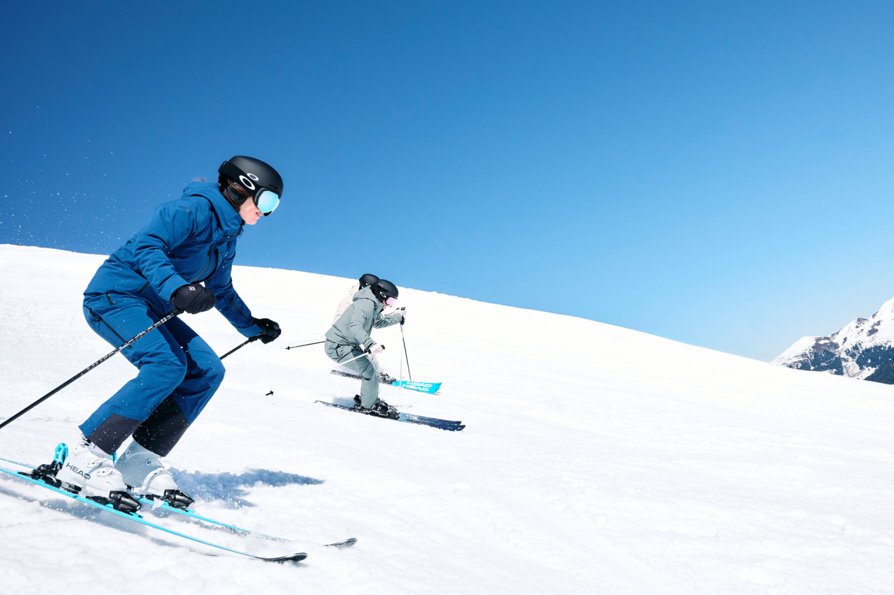 Zwei Skifahrer mit Helmen und Winterausrüstung gleiten unter einem strahlend blauen Himmel sanft einen sonnenbeschienenen Schneehang hinunter und vermitteln dabei ein Gefühl von Abenteuer und Muße.