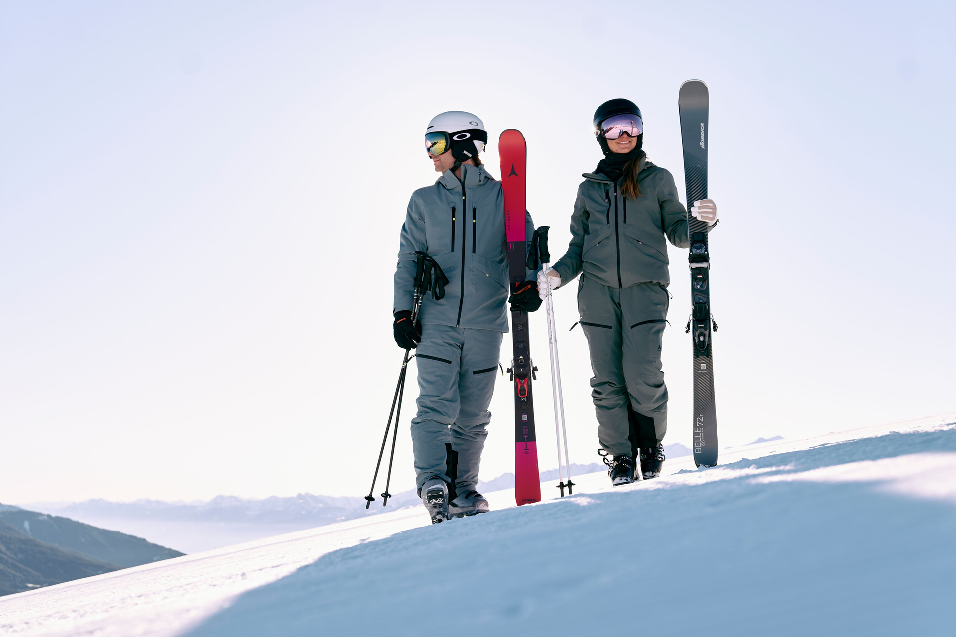 Two people in ski gear walk uphill on a snowy mountain, holding skis and poles, under a clear blue sky. The scene is calm and adventurous.