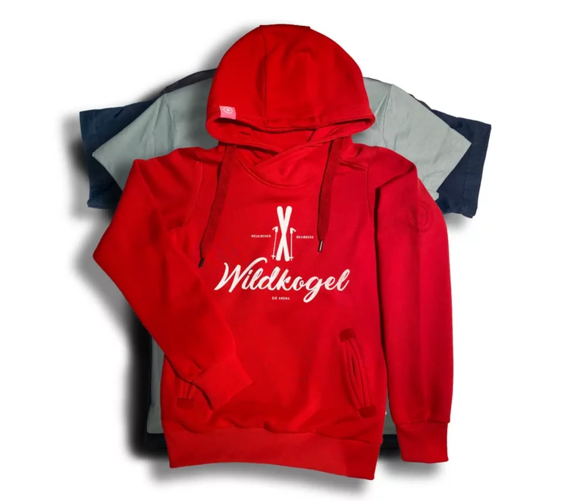Red hoodie with "Wildkogel" text and ski graphic, laid over two folded shirts in gray and blue. Casual, sporty vibe against a white background.
