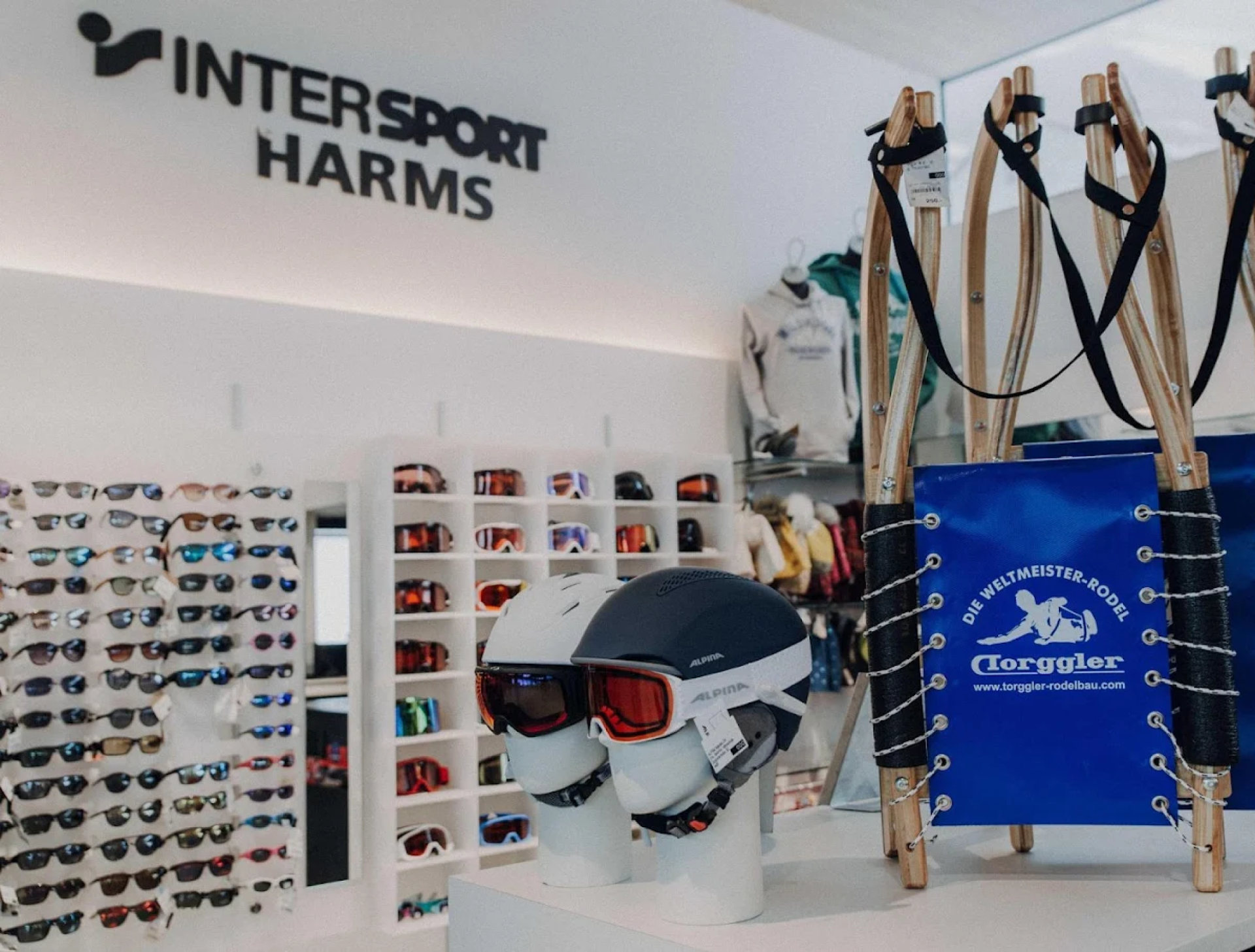 Innenraum eines Skigeschäfts mit ausgestellten Helmen und Skibrillen neben einem Holzschlitten. Im Hintergrund Regale mit Sonnenbrillen. Helle, sportliche Atmosphäre.
