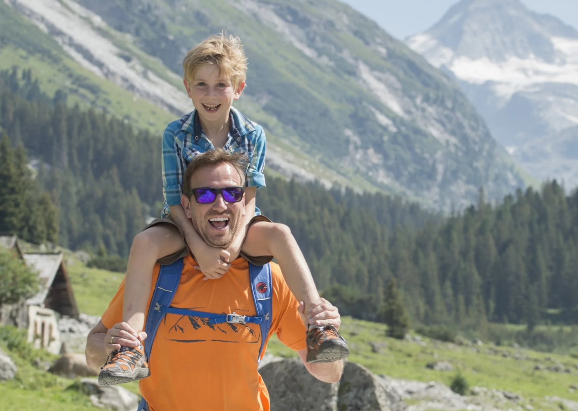 Ein Mann in einem orangefarbenen Hemd trägt freudig einen Jungen auf seinen Schultern. Sie befinden sich in einer bergigen Landschaft mit üppigem Grün und einem klaren blauen Himmel.