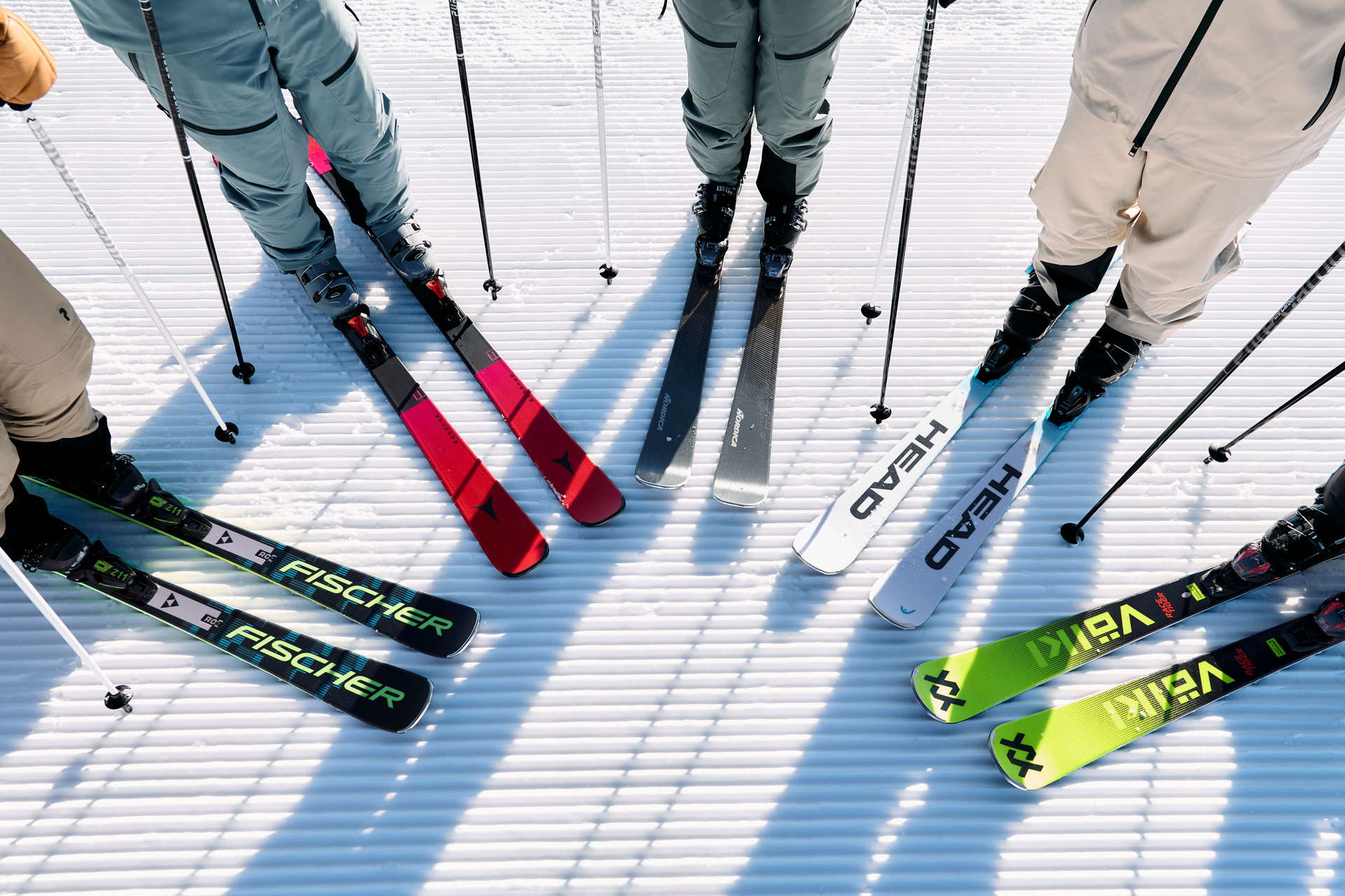 A group of skiers stands on a groomed snowy slope. Skis in various colors form a semi-circle, casting crisp shadows. The scene conveys anticipation and adventure.