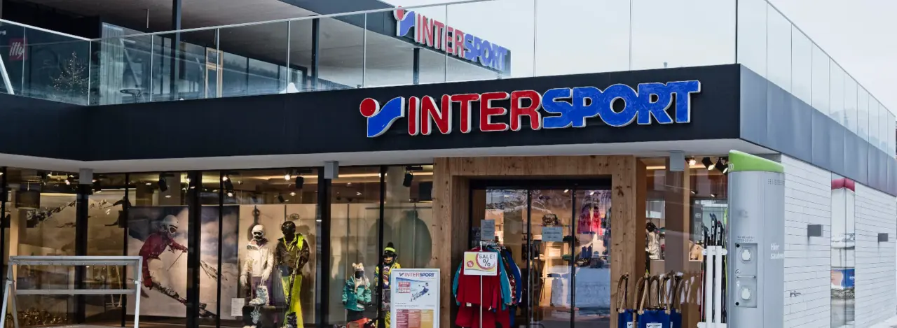 Moderne Ladenfront eines Intersport-Geschäfts mit Glasfassade. Im Schaufenster sind farbenfrohe Sportbekleidung ausgestellt, die eine einladende und energiegeladene Atmosphäre schaffen.