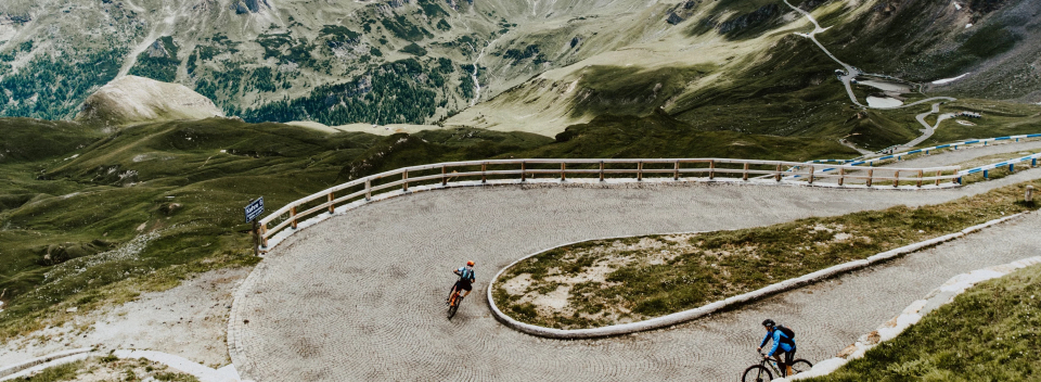Cyclists navigate a sharp mountain road curve with lush green hills and rugged terrain. The scene conveys adventure and challenge in nature.
