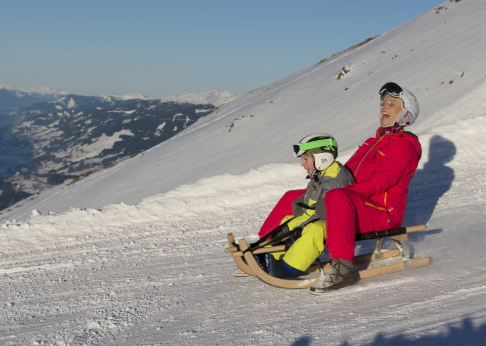 Eine Person und ein Kind in bunten Winterkleidern fahren fröhlich mit dem Schlitten einen verschneiten Berg hinunter, umgeben von fernen Gipfeln unter einem klaren blauen Himmel.