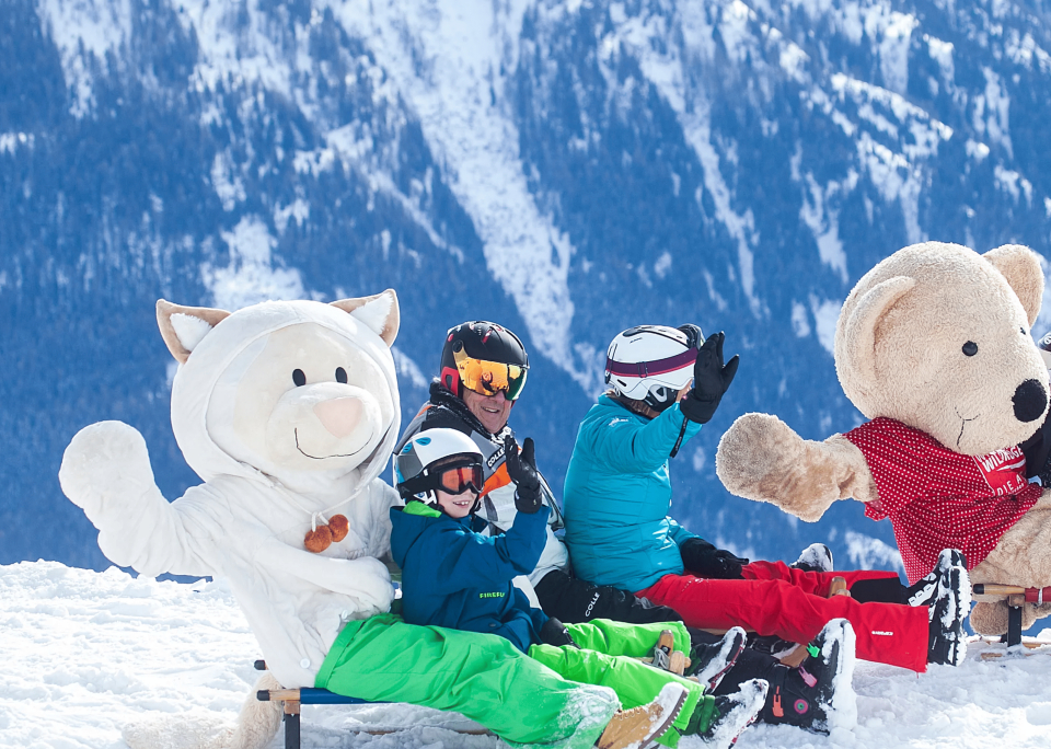 Menschen und zwei Tiermaskottchen in Schneekleidung sitzen auf einem schneebedeckten Berg, lächeln und winken. Die schneebedeckten Gipfel im Hintergrund schaffen eine fröhliche Winterszene.