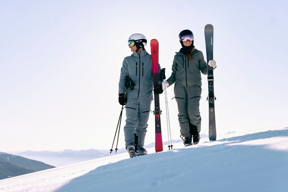 Zwei Personen in Skikleidung gehen unter einem klaren blauen Himmel mit Skiern und Stöcken in der Hand einen verschneiten Berg hinauf. Die Szene wirkt ruhig und abenteuerlich.