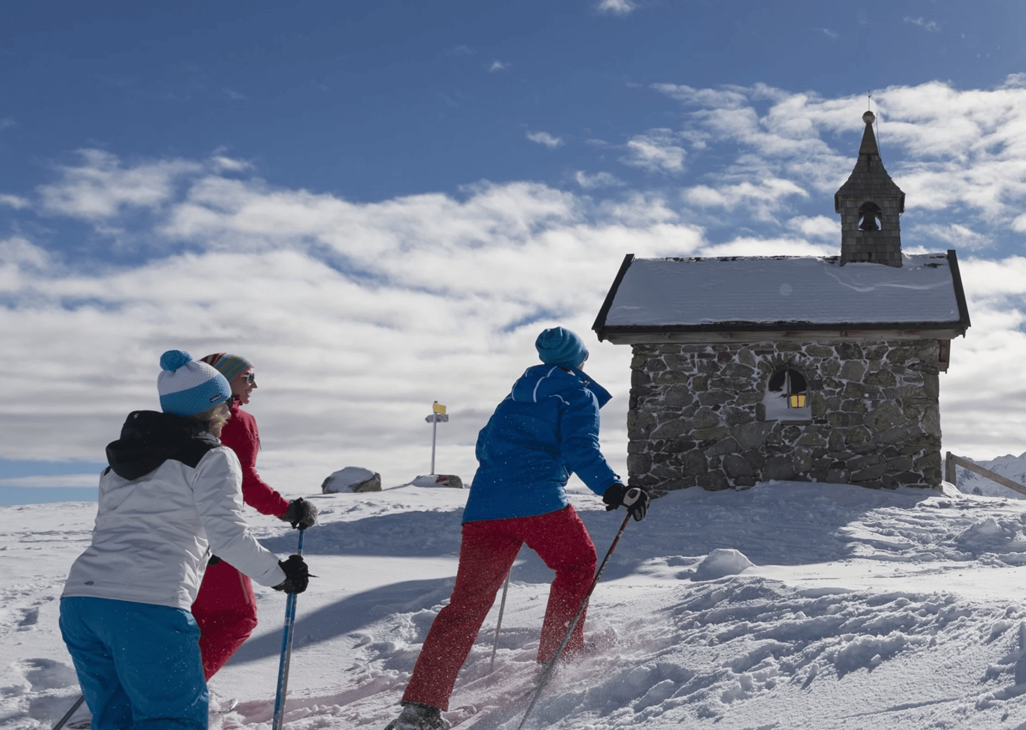 Three people in colorful winter gear ski uphill towards a small stone chapel in a snowy landscape, under a bright blue sky with fluffy clouds.