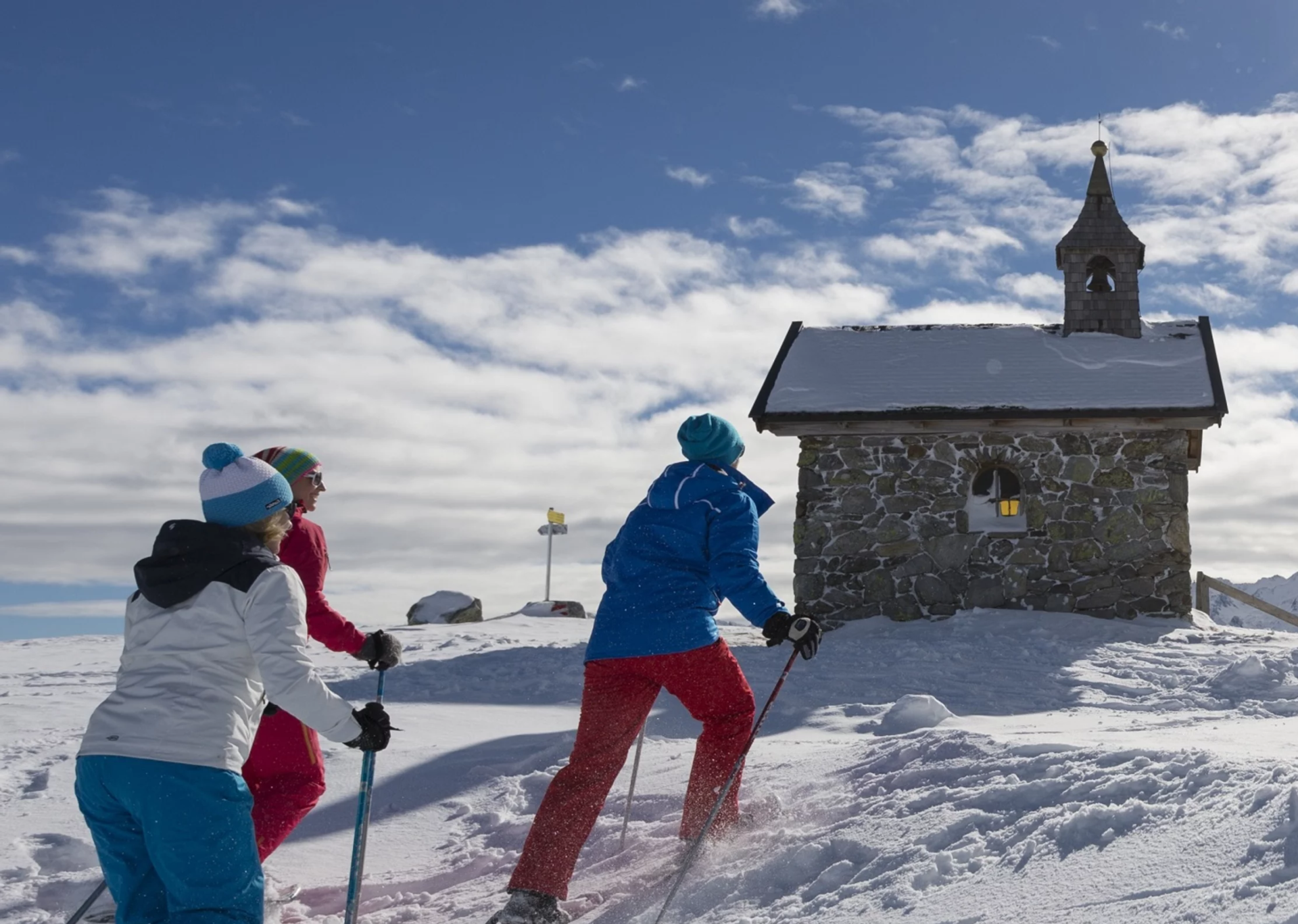 Three people in colorful winter gear ski uphill towards a small stone chapel in a snowy landscape, under a bright blue sky with fluffy clouds.