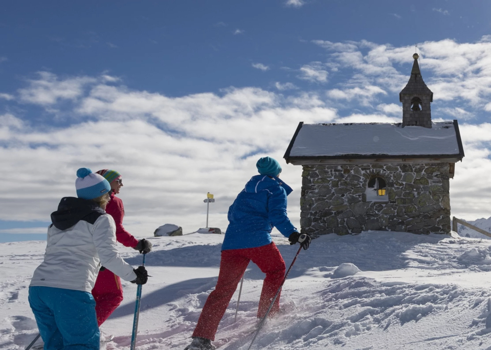 Drei Personen in farbenfroher Winterkleidung fahren mit Skiern bergauf in Richtung einer kleinen Steinkapelle in einer verschneiten Landschaft, unter einem strahlend blauen Himmel mit flauschigen Wolken.