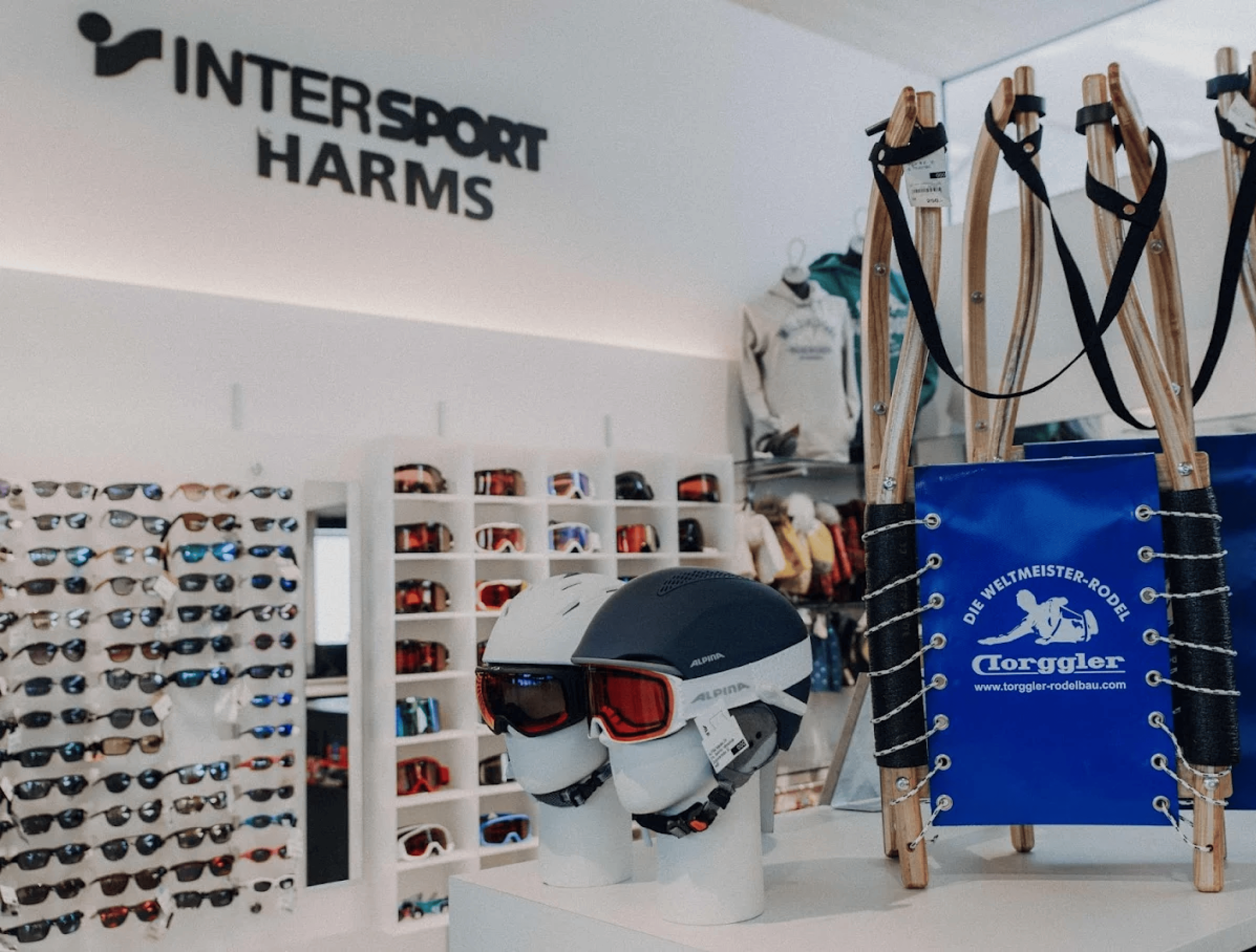 Interior of a ski shop with helmets and ski goggles on display next to a wooden sled. In the background, shelves with sunglasses. Bright, sporty atmosphere.