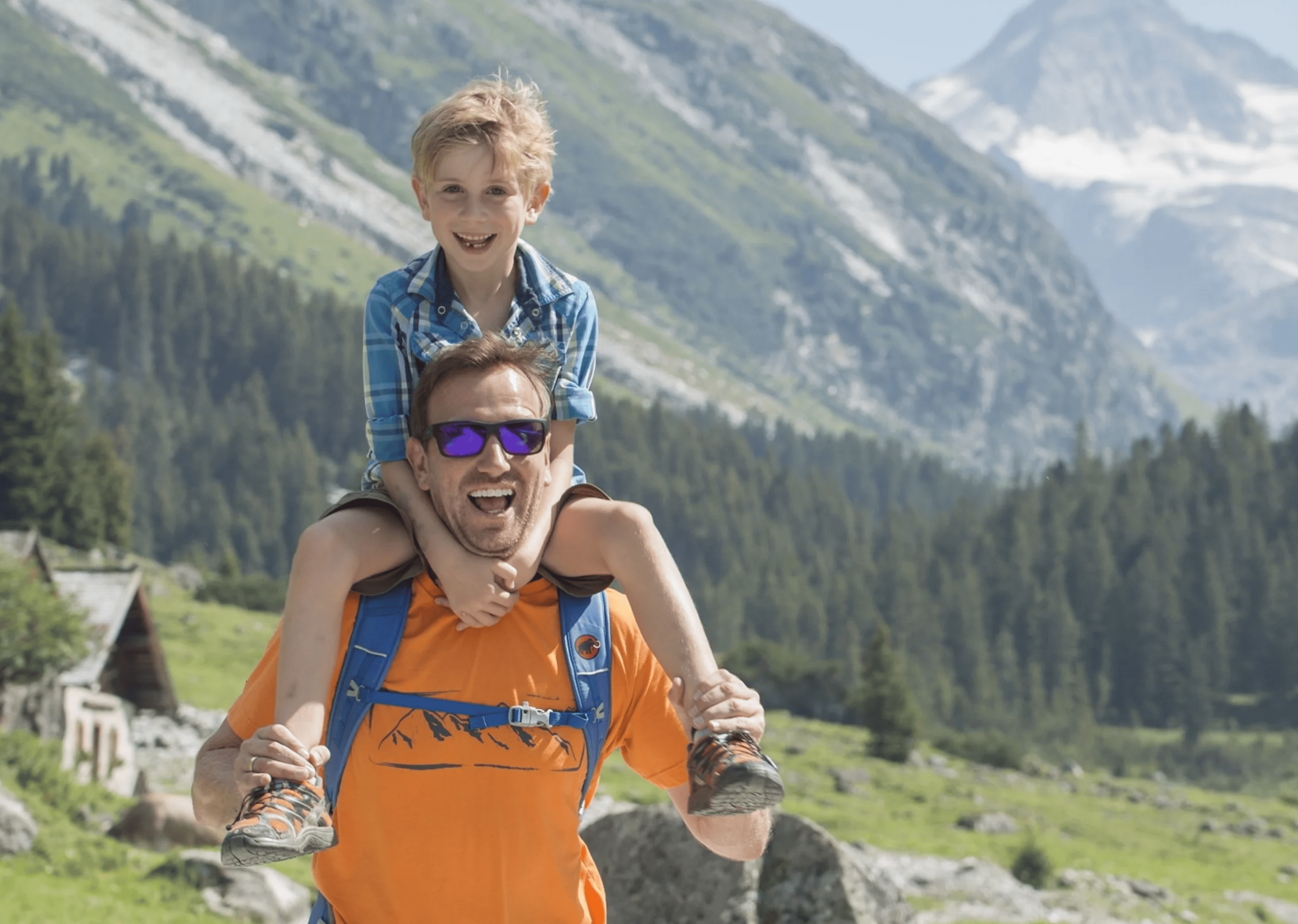 Ein Mann in einem orangefarbenen Hemd trägt freudig einen Jungen auf seinen Schultern. Sie befinden sich in einer bergigen Landschaft mit üppigem Grün und einem klaren blauen Himmel.