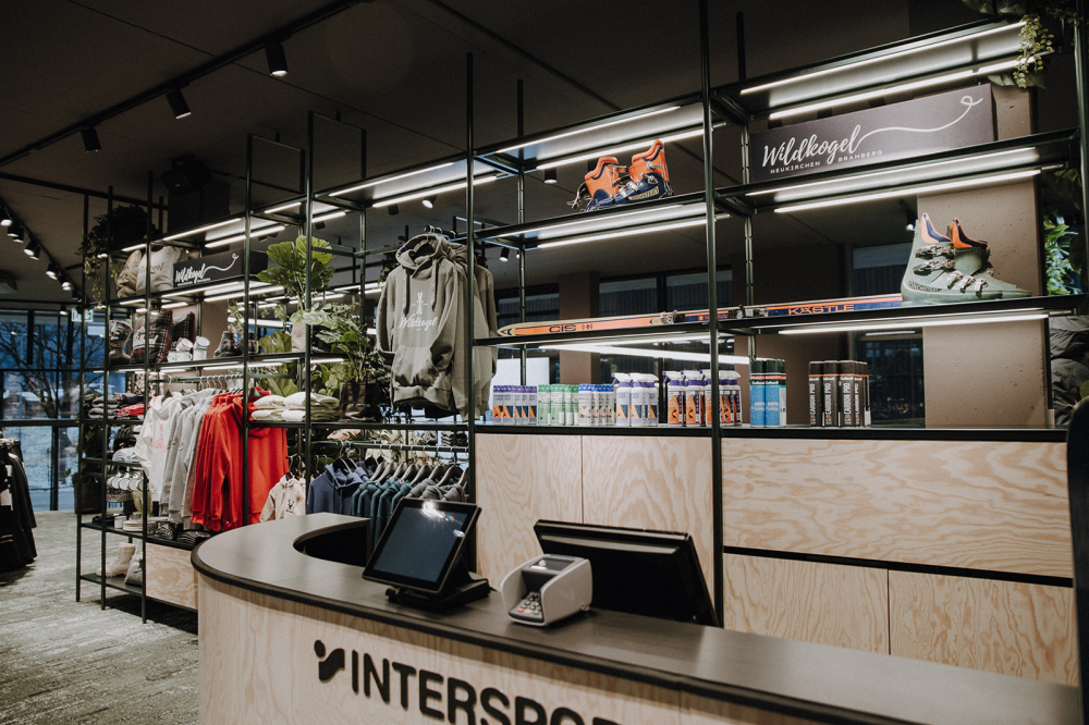 Innenraum eines Intersport-Geschäfts mit Skischuhen, Bekleidung und Ausrüstung, die auf Regalen in der Nähe der Kasse ausgestellt sind.