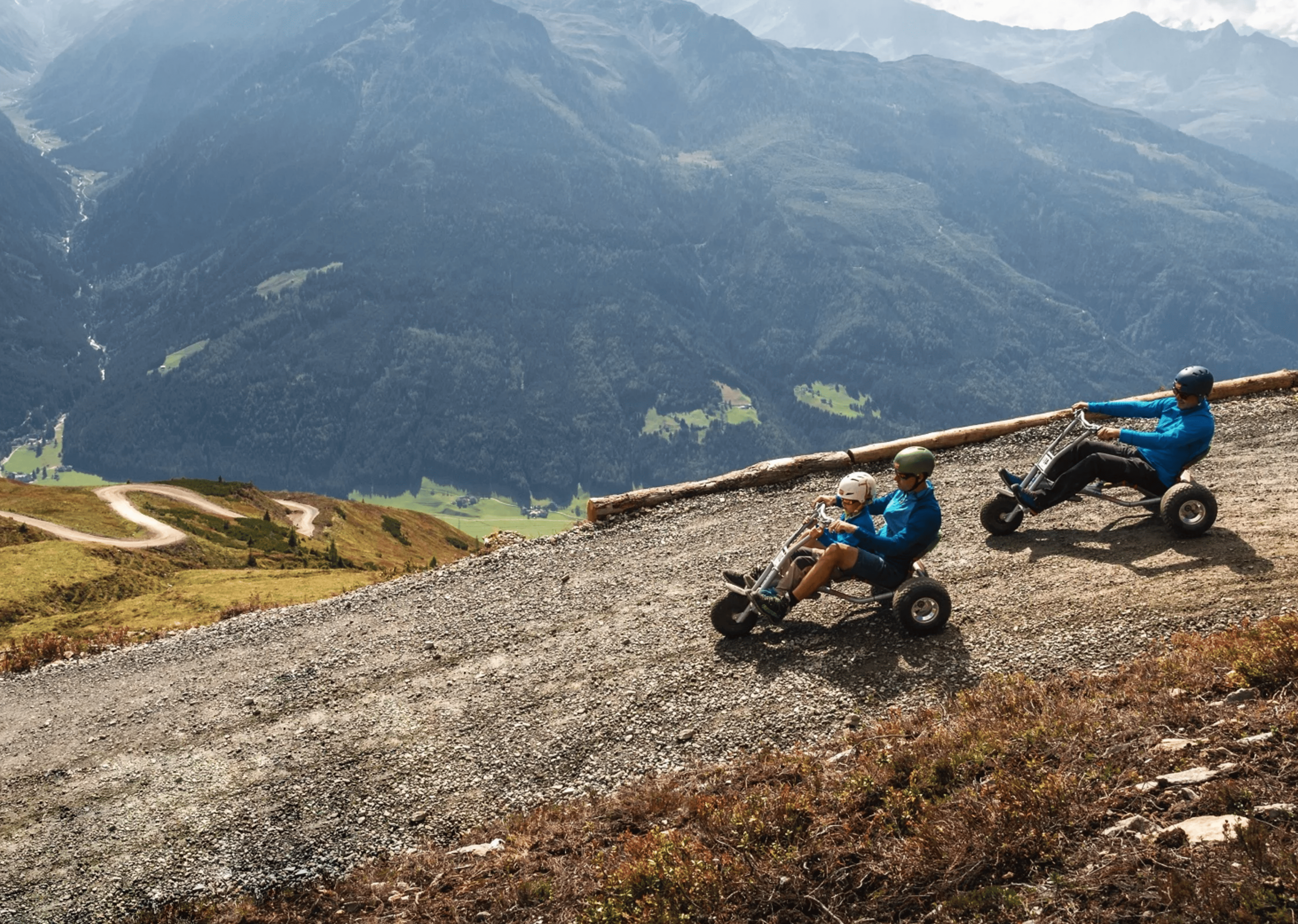 Zwei Personen mit Helmen fahren mit Tretkarts einen Bergpfad hinunter. Die malerische Aussicht umfasst grüne Hügel und neblige Berge unter einem klaren Himmel.