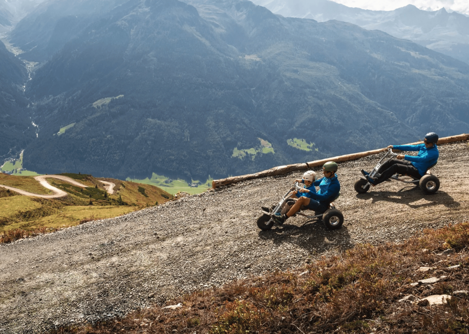 Two people wearing helmets drive pedal carts down a mountain trail. The scenic view includes green hills and misty mountains under a clear sky.