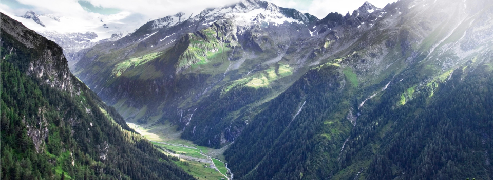 Panoramablick auf neblige Berge mit schneebedeckten Gipfeln, üppig grünen Hängen und einem hellen Tal darunter, der eine ruhige und majestätische Atmosphäre hervorruft.